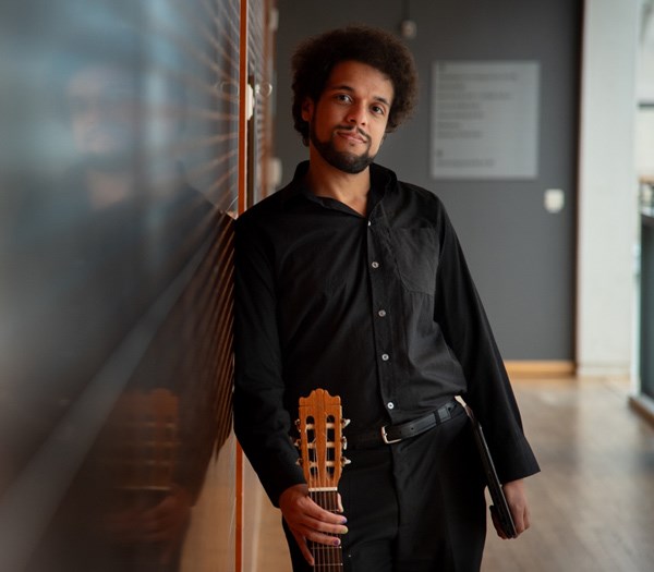 A person leaning against a wall and holding a guitar, with a reflection on the wall.