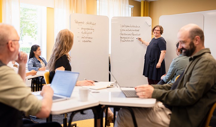 A group of men and women are listening to a woman standing by a whiteboard.