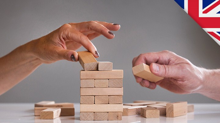 A female and a male hand build a tower of bricks. There is a British eagle flag in the right corner.