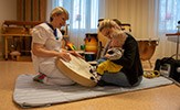 Alexandra Ullsten, i vita vårdkläder, håller fram en trumma som 1,5-årige Nils kan sparka på så att det blir ljud. De sitter på en madrass på golvet i ett rum med instrument längs väggarna.