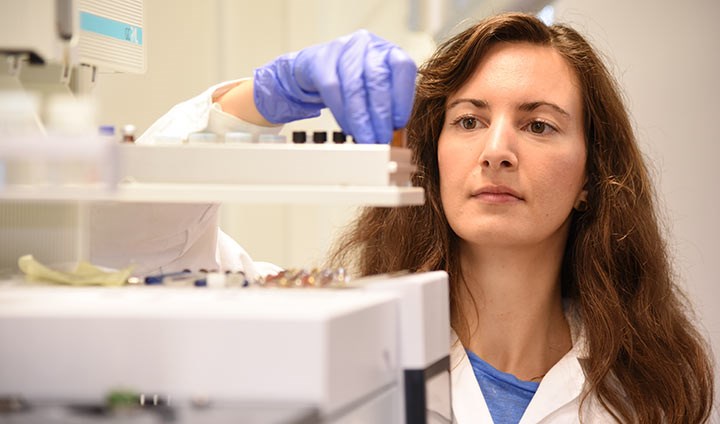 Christine Schönlau is standing in a laboratory, wearing a white coat and blue plastic gloves, holding a test tube.