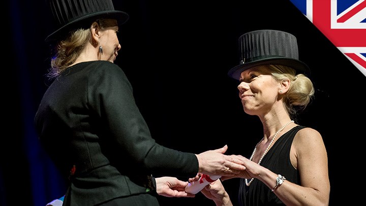 Two women in black clothes and black doctor's hats are holding hands. They look at each other.