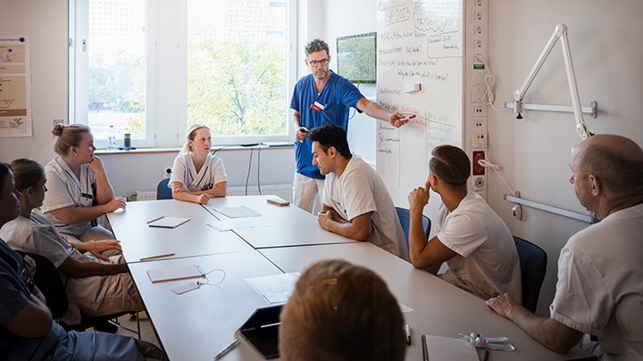 Studenter arbetar vid ett bord. En lärare står vid en whiteboard.