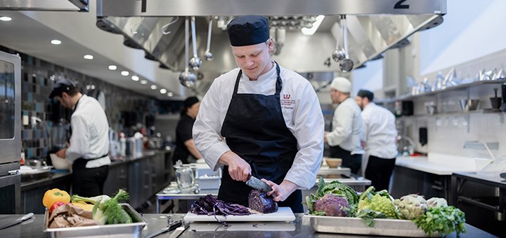 En student i kockkläder står i ett kök och hackar grönsaker.