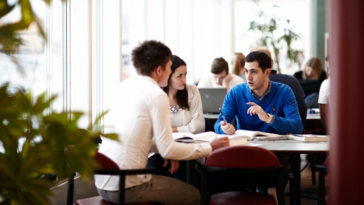 Foto på tre studenter som sitter vid ett bord med böcker och diskuterar.