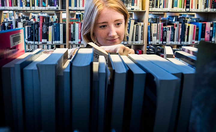 A woman stands between bookshelves in a library. Her face can be seen between the bookshelves. 