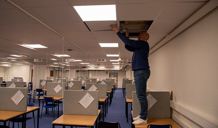 Jörgen Winbladh står på ett av många bord i en av tentasalarna. Han har lyft bort bitar av innertaket och tittar på den tåliga skyddsrumsbelysningen som finns monterad och klar under takplattorna.