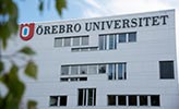 Vit byggnad med texten Örebro universitet. I förgrunden syns gröna löv.