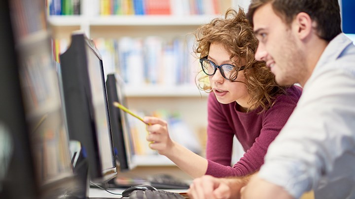 Two people sitting in front of a computer, and one of them is pointing at the screen.