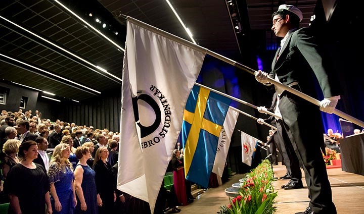 Studenter håller fanor på akademisk högtid. Finklädda människor står upp i en aula och tittar på.