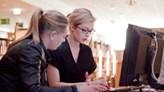 Two female students in front of a computer screen at the University Library are looking intently down at notes.