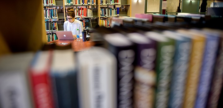 A photo of a person sitting with a laptop surrounded by bookshelves.