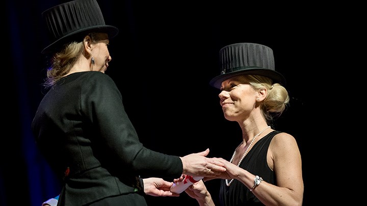 Two women in black clothes and black doctor's hats are holding hands. They look at each other.