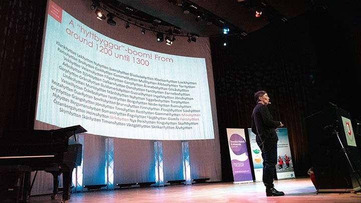 Birgitta Johansen på scen i Konsertsalen. Hon vänder sig åt höger. Bakom henne syns en bild med mängder av ortnamn som alla slutar på hytta.